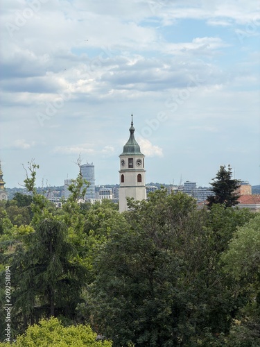 Historic Church Bell Tower Against Modern City Skyscrapers and Green Trees