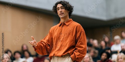 Young man addressing a seated audience in a modern auditorium background for speech presentation and event banner