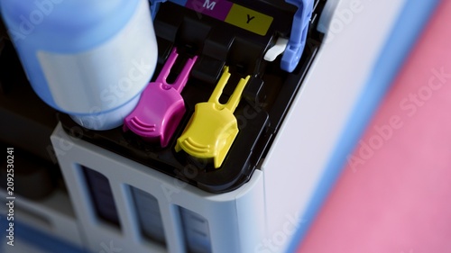 Photos A close-up of filling an inkjet printer tank system with a cyan ink bottle next to magenta and yellow color ports with plastic plugs