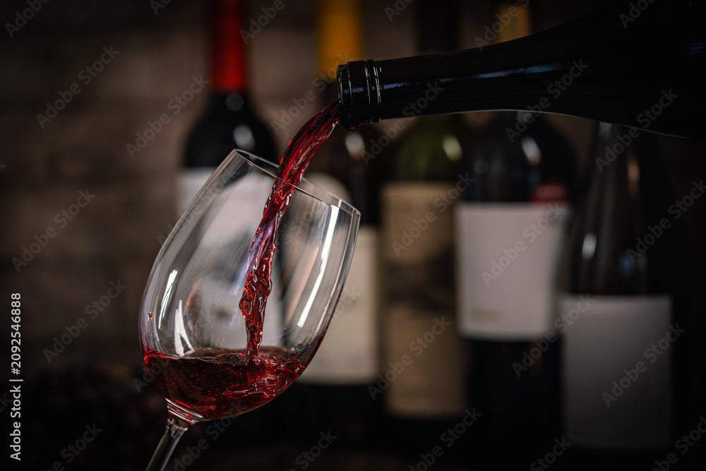 Obraz premium A dark, elegant still life of red wine being poured into a glass with wine bottles and fresh grapes in the background. Perfect for wine-themed compositions