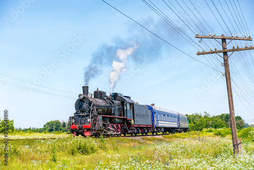 A vintage steam locomotive pulling passenger cars through a scenic green field under a clear blue sky, emitting dark smoke and white steam near a wooden utility pole.
