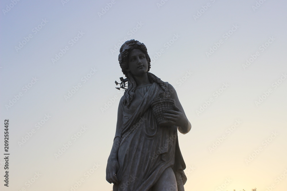 Fototapeta premium Stone statue of woman carrying harvest basket against soft pale twilight sky background.