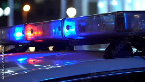 Close-up emergency vehicle lightbar glowing with red and blue LEDs on glossy rooftop at night, casting vivid reflections across dark surfaces and showing urgent warning light detail