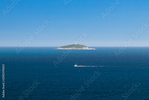Obraz na plátne A view of several boats sailing on the blue sea on a sunny day; in the distance, a small, rocky, uninhabited island is visible, though it has small trees at the top