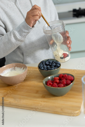 Yogurt and berries are being stirred in clear mixing cup using wooden spoon on kitchen countertop