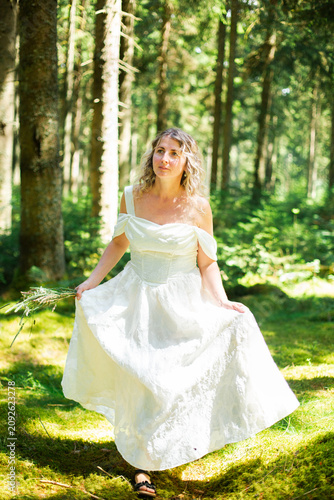 Fotografie Smiling blonde woman in a long white off-shoulder dress walks toward the camera while holding her flowing skirt in a sunlit green forest