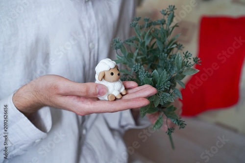Vintage white sheep symbol of 2027 figurine in the hands of a young man on a background of white linen shirt and green mistletoe branches close-up