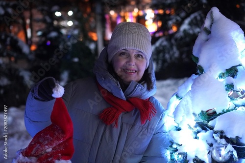 Smiling woman in winter clothing holds a Santa hat beside a lit snow-covered Christmas tree. The nighttime scene captures the festive atmosphere and joyful spirit