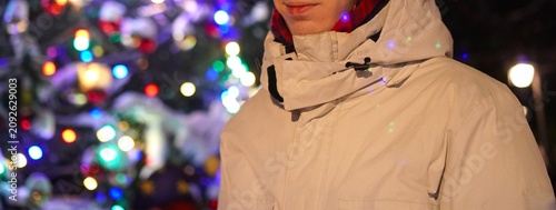 Outdoor scene with person in winter attire, enjoying a festive moment beside a decorated Christmas tree with snow, glowing lights, and joyful ambiance, capturing holiday cheer.