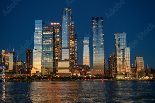 New York, USA, 8 June 2026 : Hudson Yards skyline, view of skyscrapers from the Hudson River. Lit office buildings and the Vessel structure reflect on dark water.