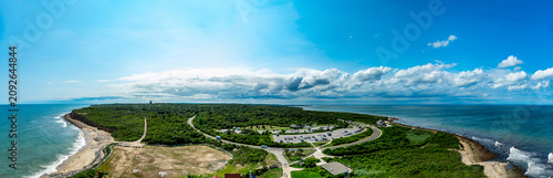 Wide panoramic view of Montauk Point coastline in New York. A green peninsula is surrounded by ocean waters with sandy beaches and roads visible from a high angle.