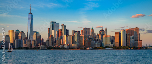 Lower Manhattan skyline with One World Trade Center at sunset. Wide panoramic view of New York skyscrapers reflecting warm evening sunlight with a sailboat on the river.