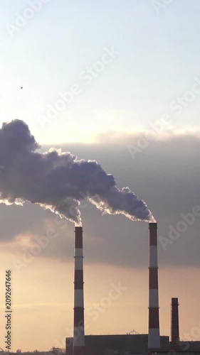 Industrial smokestacks release thick smoke into a cloudy backlit sky as emissions drift over a factory at sunset, highlighting air pollution and heavy industry