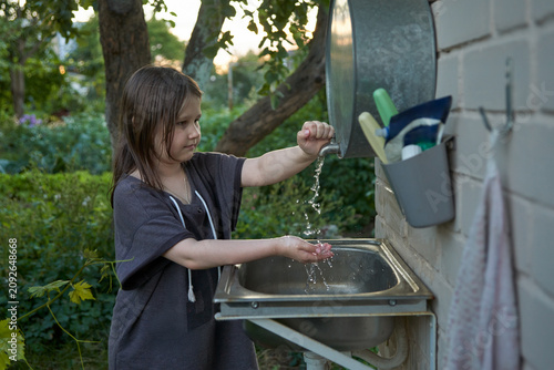 Canvas Print A little girl washes her hands in a washstand attached to the wall of a brick building at a summer house