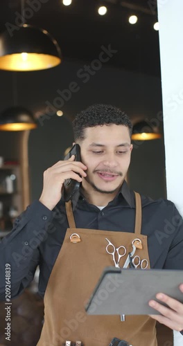 Vertical video: African American man barber in shop apron on phone using tablet to confirm slots