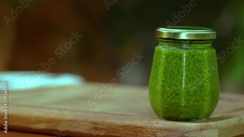 A jar of pesto on a wooden board with a blurred background. Pesto sauce, green basil.