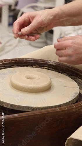 High-angle close-up artisan hands holding tool above centered wet clay disk on spinning pottery wheel within wooden splash pan, representing creative handmade ceramic craft workshop.