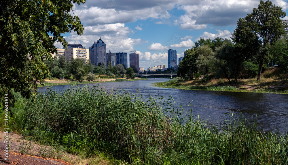 Fototapeta premium Cityscape showcasing modern apartment buildings reflecting urban development, coexisting with a wide winding river, lush green trees, and natural vegetation under a cloudy sky
