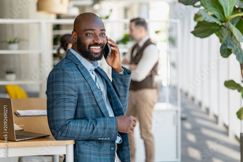 Smiling businessman on phone in modern office leaning on desk with laptop and plants