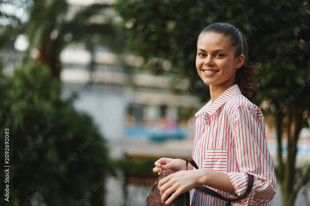 Naklejka premium Cheerful woman outdoors in striped shirt, smiling, natural light, soft breeze, casual pose amid greenery and relaxed moment.