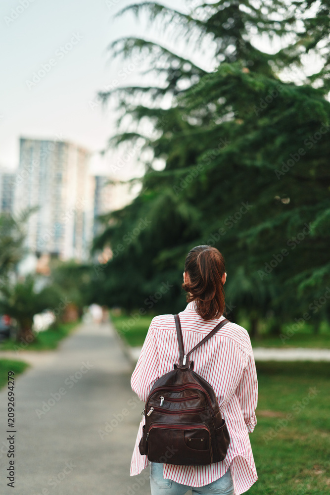 Naklejka premium Woman with backpack walking along a park path, casual outfit in an outdoor setting, capturing lifestyle and urban nature in a candid moment, sunny mood