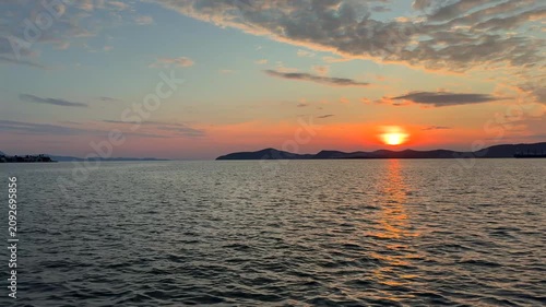 A calm, warm sunset over the Aegean Sea in Gulluk, Turkey. The sun hangs low in a hazy orange sky, casting a bright golden light path across the gently rippling water toward distant dark hills.