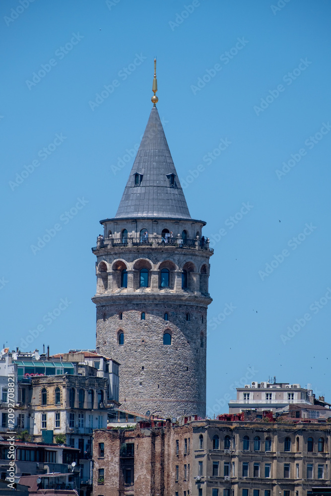 Obraz premium Galata Tower Rising Between Historic Buildings in Istanbul. Historic Galata Tower Surrounded by Istanbul Buildings. Galata Tower Emerging Above the Cityscape.