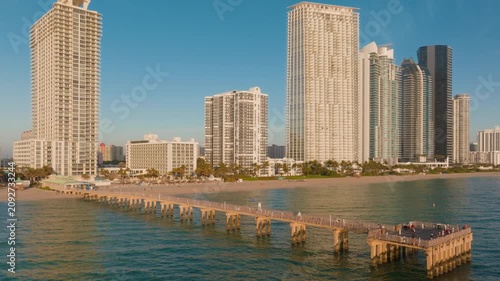 Aerial drone footage of Sunny Isles Beach fishing pier extending into turquoise Atlantic Ocean waters with luxury highrise condominiums and beachfront hotels lining the sandy Miami coastline.