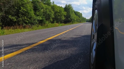 Rural countryside highway asphalt road drive. Cinematic POV of the front left car wheel driving plate. Yellow line lane separator in the frame. Cottage country commute, back of wheel exterior view.