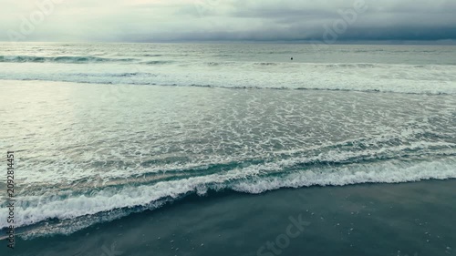 Aerial drone view of rolling ocean waves across Pacific Beach shoreline beneath soft cloudy California coastal skies above