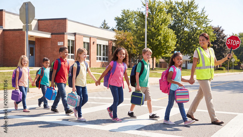 School crossing guard holds a stop sign while guiding a line of young elementary students across the crosswalk to reach their school building safely.