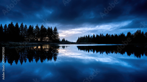 A serene lake reflecting the dramatic, cloudy sky and silhouetted pine trees during twilight. The dark blue hues create a moody and tranquil atmosphere