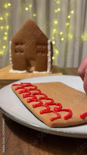 Vertical 4K video close up. 60 fps. Female hand with plastic bag with edible paint draws red wave pattern on brown roof of gingerbread house in plate on table. Sweet food for winter holidays, New Year