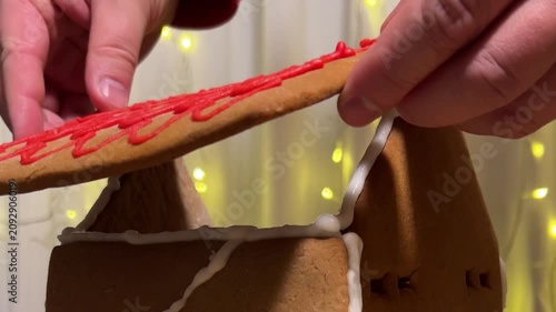 Close up video. 60 fps. Male hands glues roof with red wave pattern, edible paint to brown gingerbread house with white icing and broken wall. Garland on curtains out of focus. Bokeh. New Year 2027