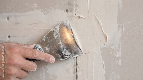 Hand holding putty knife applying filler compound over drywall seams during home renovation with construction renovation finishing
