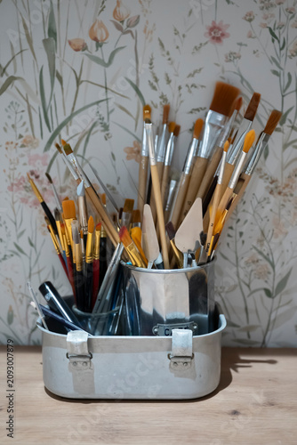 Various painting brushes with wooden handles arranged in metal containers inside an artist’s studio, representing creativity, art supplies and traditional painting techniques.