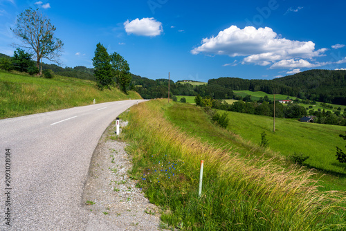 A winding country road curves through lush green rolling hills in the picturesque Austrian countryside. Bright blue sky with fluffy white clouds stretches above distant forests and quiet farmland