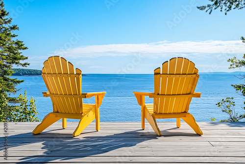 Two Adirondack chairs facing the ocean beneath a bright sunny coastal sky.