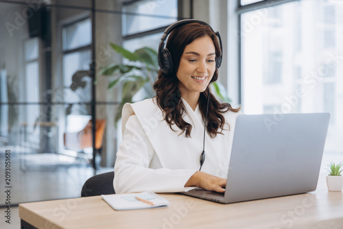Young businesswoman wearing headphones and smiling, typing on laptop while working in a modern office
