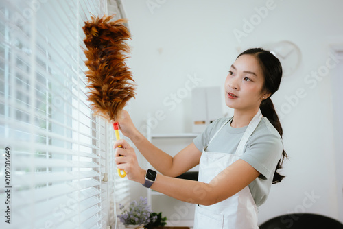 Canvastavla Young woman dusting window blinds with a feather duster in a bright modern living room, keeping her home clean and fresh