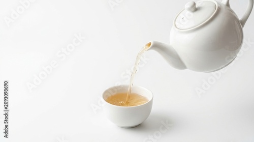 Side angle view of an elegant round white porcelain kettle pouring organic herbal tea into a cup