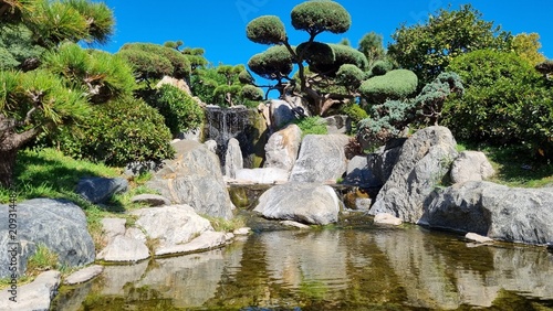 Scenic oriental garden featuring trimmed bonsai trees, decorative pond, and stone waterfall under blue sky.