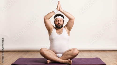 Overweight man with a beard in a white tank top and briefs doing a yoga pose on a purple mat, hands in prayer position above his head.