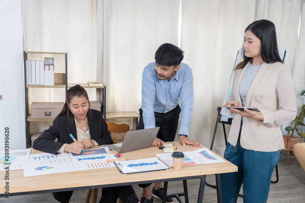 Fototapeta premium Asian Business Team Working on Project: Three Asian professionals actively work on documents and digital devices, discussing a project in a bright, modern office. Highlights focused collaboration.