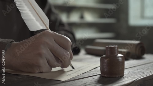 Monastic scribe in a brown robe dipping a large quill pen into a clay inkpot and writing a manuscript on parchment at a wooden table, with scrolls in the blurred background