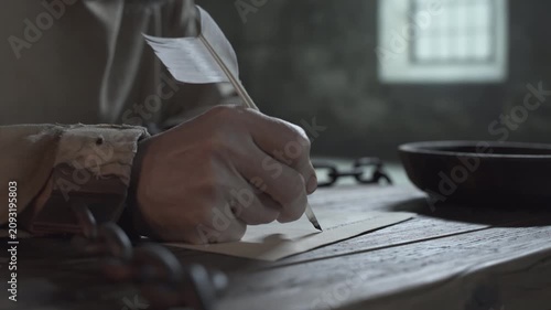 Close up of medieval prisoner chained hands writing the initial line with a quill pen on parchment paper on a rustic wooden table inside a gloomy dungeon 