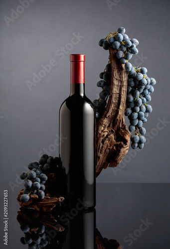 Bottle of red wine with a weathered snag and blue grapes on a black reflective background.
