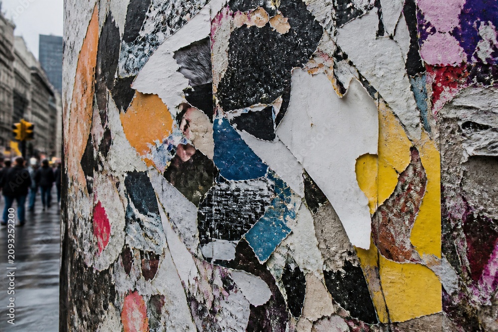 Naklejka premium A close view of torn paper and colorful posters on a column. City streets are wet from rain, and people are seen walking in the distance.