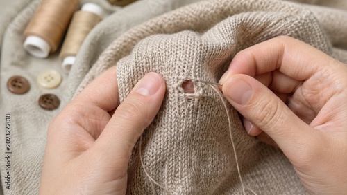 Hands darning a hole in a beige knitted sweater using needle and thread