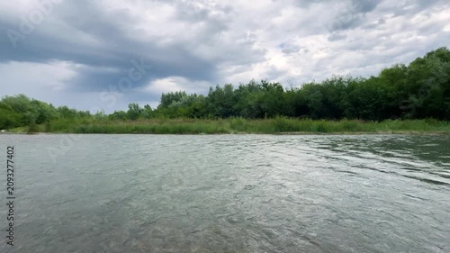 A calm river flows past green grassy banks under a cloudy sky. Dense summer trees grow along the opposite shoreline in daylight as water surface ripples gently through this natural landscape.